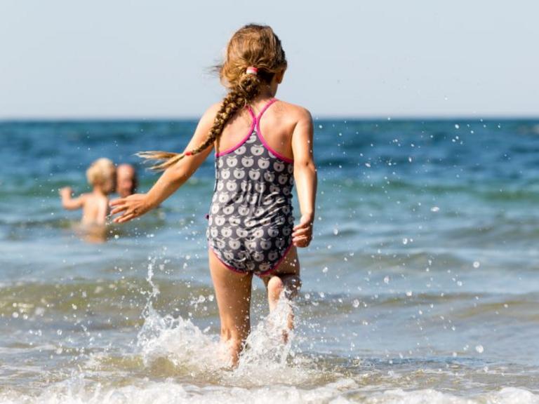 Pige løber ud i vandet ved stranden, mens andre børn bader