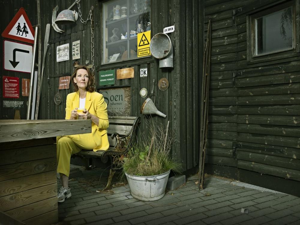 Ditte Hansen i gult jakkesæt på bænk med kaffekop og mange skilte i baggrunden 