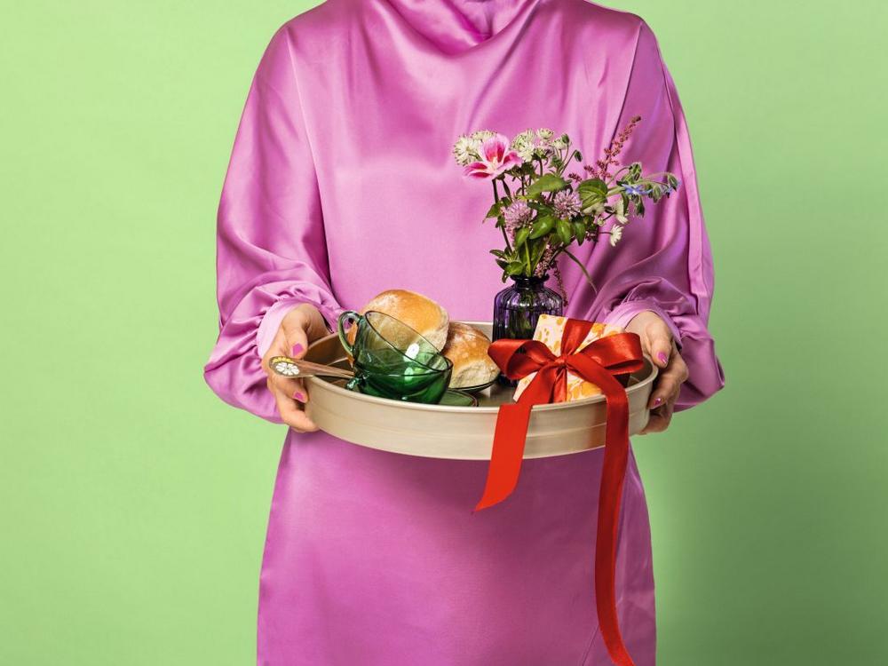 Person i lyserød kjole på grøn baggrund holder bakke med boller, blomster og en gave